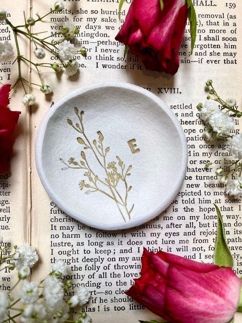 May include: A white ceramic ring dish with a gold floral design and the letter "E" in the center. The dish is sitting on a page of a book with text.