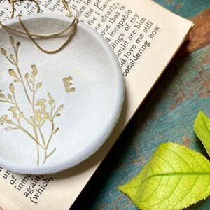 May include: A white ceramic ring dish with a gold floral design and the letter "E" in the center. The dish is sitting on a book with a gold chain necklace on top of it.