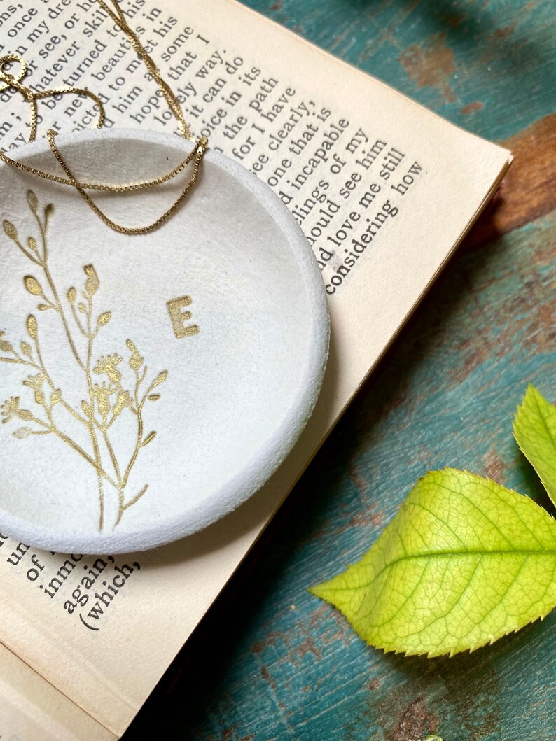 May include: A white ceramic ring dish with a gold floral design and the letter "E" in the center. The dish is sitting on a book with a gold chain necklace on top of it.