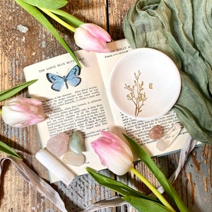 May include: A white ceramic trinket dish with a gold floral design and the letter "E" in the center. The dish is surrounded by pink tulips, green leaves, and various crystals.
