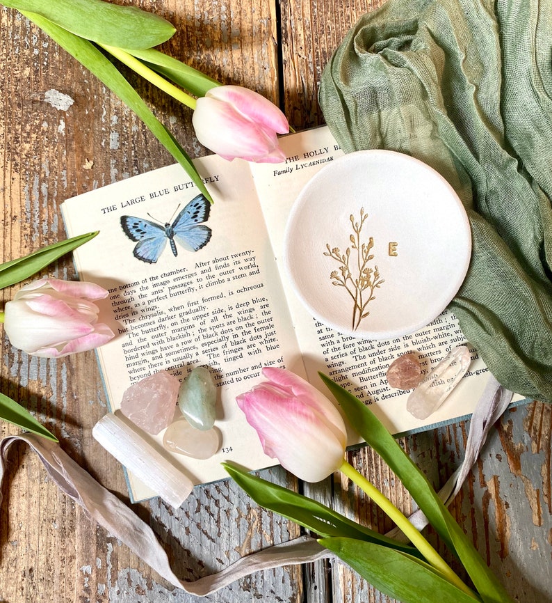 May include: A white ceramic trinket dish with a gold floral design and the letter "E" in the center. The dish is surrounded by pink tulips, green leaves, and various crystals.