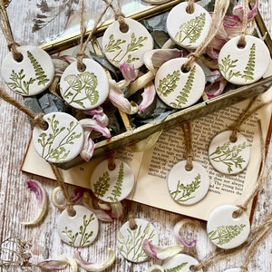 May include: A collection of 14 white ceramic ornaments with green floral designs. The ornaments are strung with twine and are arranged on a wooden surface with dried flower petals.