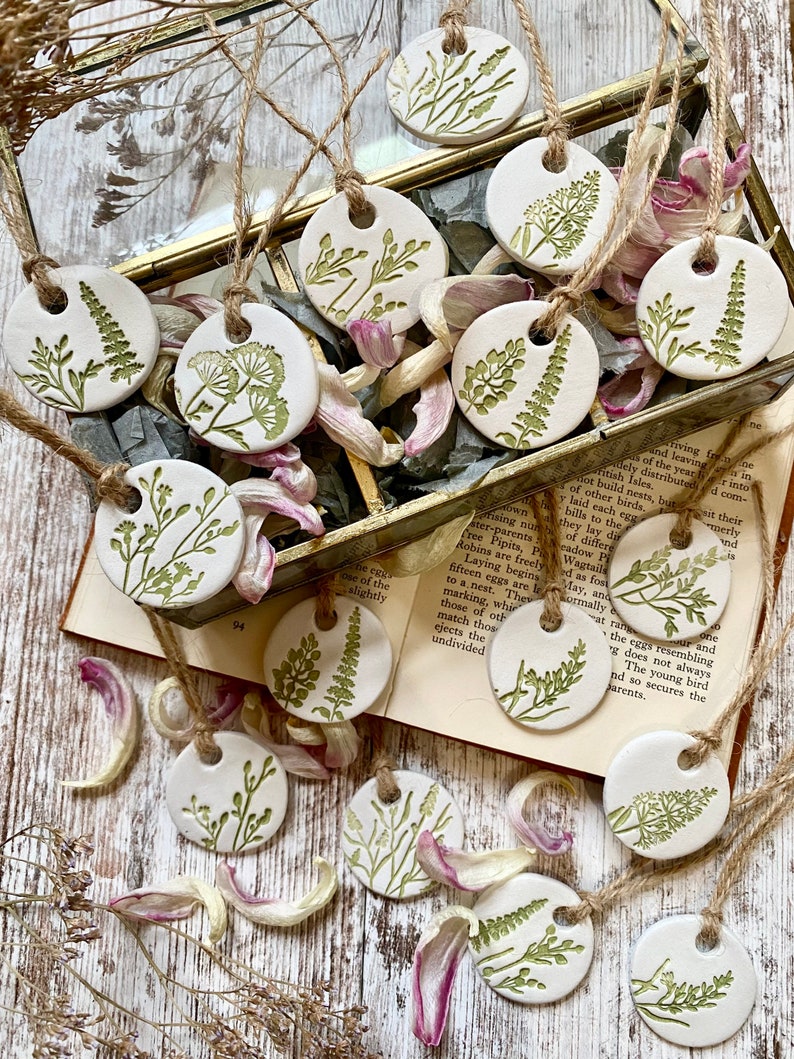 May include: A collection of 14 white ceramic ornaments with green floral designs. The ornaments are strung with twine and are arranged on a wooden surface with dried flower petals.