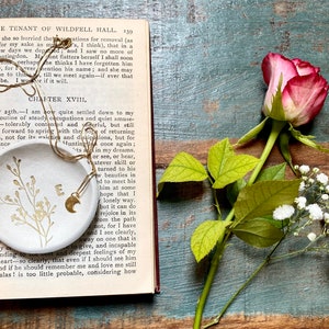 May include: A white ceramic dish with a gold chain and a crescent moon charm. The dish has a delicate floral design and the letter "E" in the center. The dish is sitting on an open book with the text "Chapter XVIII" visible.