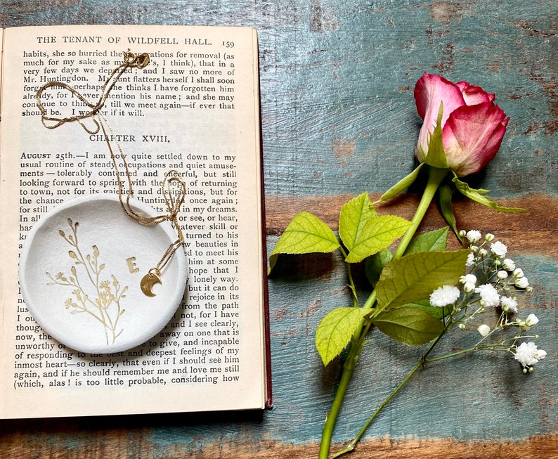 May include: A white ceramic dish with a gold chain and a crescent moon charm. The dish has a delicate floral design and the letter "E" in the center. The dish is sitting on an open book with the text "Chapter XVIII" visible.