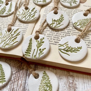 May include: A set of 10 white ceramic ornaments with green floral designs. Each ornament has a hole at the top for hanging. The ornaments are arranged on a book with text about birds.