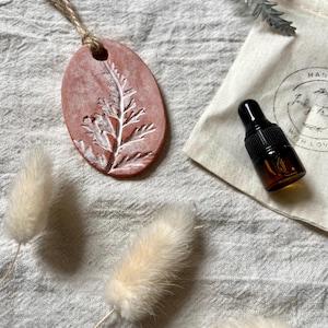 May include: A terracotta-coloured ceramic ornament with a white fern design, hanging from a twine string. The ornament is on a white fabric background with a small bottle of essential oil and a white pouch.