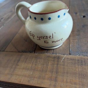 May include: A cream-colored ceramic creamer pitcher featuring a handle, a spout, and a brown rim. The exterior is decorated with a row of small blue dots near the top and the hand-painted black text that reads, 'Elp yersel' to more'.