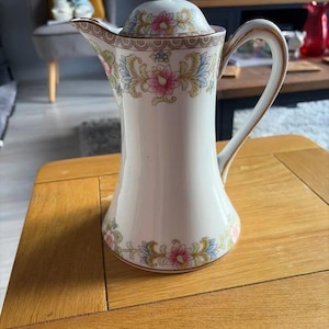 Op de afbeelding: Een witte porseleinen koffiepot met een delicaat bloemmotief in roze, blauwe en groene tinten. De pot is voorzien van een bijpassend deksel met knop, een gebogen handvat en een uitlopende tuit, alles afgewerkt met een fijne gouden rand.