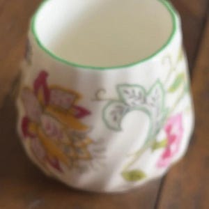May include: A small, white ceramic cup with a fluted, rounded body. The exterior features a colourful floral pattern with pink, yellow, and green botanical motifs. The rim of the cup is accented with a thin, solid green painted line.