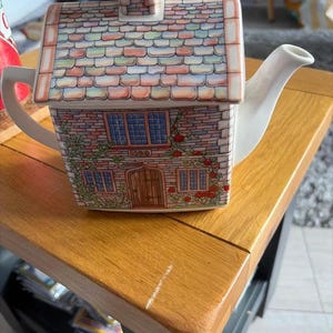 Può includere: Una teiera in ceramica a forma di cottage con esterno in mattoni, camino e tetto in tegole. La casa presenta una porta in legno, diverse finestre e rose rampicanti. Un manico e un beccuccio bianchi sporgono dai lati della struttura.