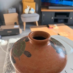 May include: A round, terracotta-coloured clay vase with a wide, bulbous body and a narrow, circular opening. The exterior surface features hand-painted green and black turtle motifs with detailed shell patterns encircling the vessel.
