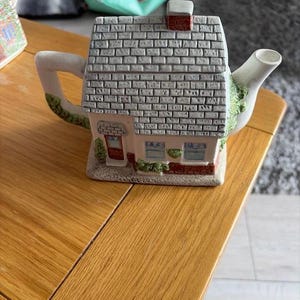 Pode incluir: Um bule de cerâmica em forma de pequena casa de campo com telhado cinzento texturizado, chaminé vermelha e paredes brancas. A estrutura apresenta uma porta castanha, janelas com molduras azuis e detalhes de folhagem verde na base e no bico.
