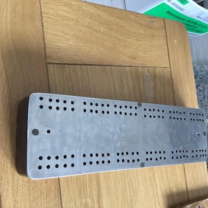 May include: A rectangular cribbage board featuring a metallic top surface with multiple rows of drilled holes for peg placement. The board has a dark wood base and is constructed with a flat, elongated design, showing visible screw heads.