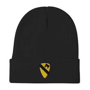 Puede incluir: Gorro negro con borde doblado. El gorro presenta un emblema bordado en amarillo de un escudo con una franja diagonal negra y una silueta de caballo. El gorro está hecho de un material suave y tejido.