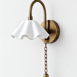 May include: A wall-mounted lamp featuring a curved antique brass arm and a circular wall plate. The light fixture includes a white, ruffled, bell-shaped shade and a hanging brass chain pull with a spherical end, providing a functional design.