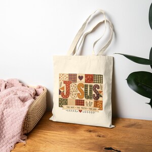 Peut inclure: Un sac fourre-tout en toile couleur crème arborant un motif central avec le mot JESUS en grandes lettres style patchwork. Le design inclut divers motifs floraux et à carreaux dans des tons chauds, avec le texte THE WAY THE TRUTH THE LIFE en dessous.