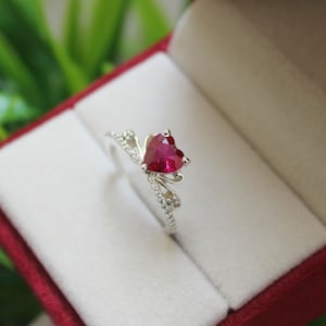 Könnte beinhalten: Ein silberner Ring mit einem herzförmigen roten Edelstein in der Mitte. Die Ringschiene teilt sich in zwei zarte, geschwungene Arme, die am Stein zusammenlaufen und beide mit kleinen, klaren, funkelnden Steinen am Rand besetzt sind.