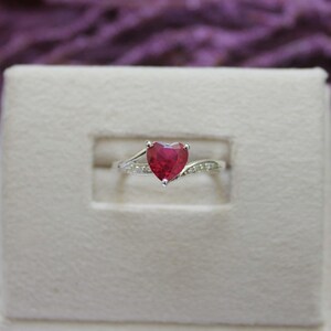 May include: A silver ring featuring a prominent heart-shaped red gemstone at the centre. The band is crafted from a polished silver-toned metal and is accented with small, clear, sparkling stones embedded along the sides of the ring shank.