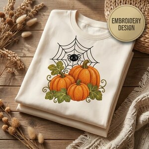 Pode incluir: Uma t-shirt cor de creme com um gráfico de três abóboras cor de laranja com folhas verdes e uma aranha preta numa teia. O texto EMBROIDERY DESIGN está impresso num círculo dourado no canto superior direito da imagem.