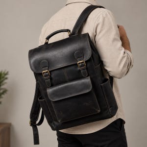 Può includere: Uno zaino in pelle nera dotato di manico superiore, due spallacci regolabili e una patta ripiegabile fissata con due fibbie in metallo color ottone. La parte anteriore presenta una grande tasca rettangolare con patta e tasche laterali.