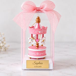 May include: A miniature carousel ornament inside a clear rectangular display box topped with a pink ribbon. The carousel features a white horse with pink accents, a pink canopy, and a gold base. A gold plaque on the box reads Sophia 12.05.2025.