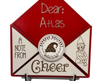 May include: A red wooden envelope-shaped sign with the words "Dear: Atlas" in silver lettering. The sign also features the text "A Note From Cheer" and an illustration of an elf. A circular emblem reads "Express Delivery Official Elf Mail."