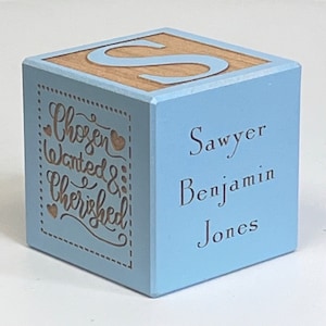 May include: A light blue wooden block with the letter "S" on one side and the words "Chosen Wanted & Cherished" on another side. The other side of the block has the name "Sawyer Benjamin Jones" engraved on it.