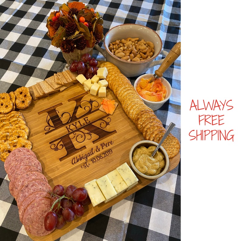 May include: Personalized wooden serving board with the names Kyle and Abigail & Pere, established 10.06.2019, engraved on the surface. The board is filled with various snacks, including crackers, cheese, salami, grapes, and dips.