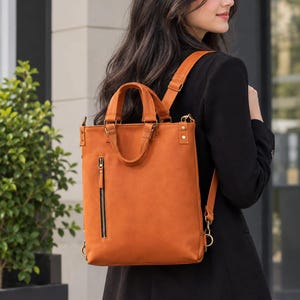 Könnte beinhalten: Ein rechteckiger orangefarbener Rucksack mit glatter, matter Oberfläche, ausgestattet mit zwei Tragegriffen oben, verstellbaren Schultergurten und einer vertikalen Reißverschlusstasche auf der Vorderseite. Die Tasche hat Metallbeschläge und seitliche Ringe.