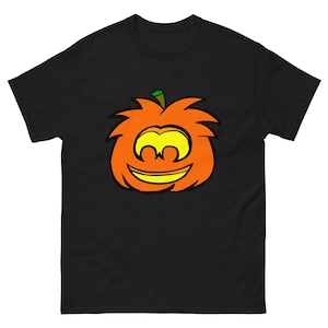 May include: A black short-sleeve t-shirt featuring a centered graphic of a stylized orange pumpkin with a green stem. The pumpkin has a yellow face with large, rounded eyes and a wide, curved mouth, all outlined in thick black lines.