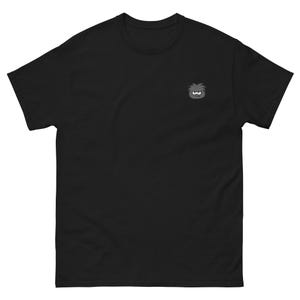 Può includere: Una t-shirt nera a maniche corte con una piccola grafica ricamata sulla parte superiore sinistra del petto. La grafica raffigura una creatura grigia, rotonda e stilizzata, con capelli spettinati e un'espressione facciale imbronciata in grigio e bianco.
