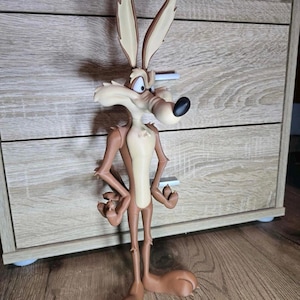 May include: A Wile E. Coyote figurine featuring a brown body with a cream-colored chest and muzzle, tall pointed ears, and a black nose. The character is depicted in a standing pose with arms bent at the elbows and hands resting on the hips.