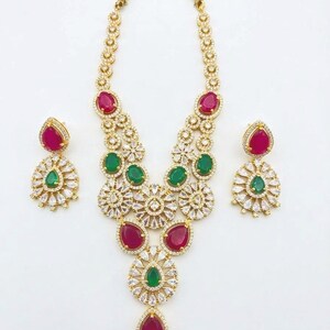 May include: A gold-tone necklace and earring set featuring teardrop-shaped red and green stones surrounded by clear pavé crystals. The necklace has a multi-layered floral design with a central pendant, and the earrings are matching studs.