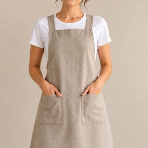 May include: A beige apron featuring a square neckline, thick shoulder straps, and two large front patch pockets. The apron is worn over a plain white short-sleeved t-shirt and blue denim jeans, providing a neutral and functional appearance.