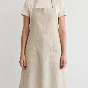May include: A beige apron featuring a square neckline, two front pockets with flaps, and adjustable neck and waist straps. The apron is crafted from a textured fabric and provides full coverage from the chest down to the knees.