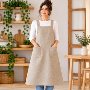 Könnte beinhalten: Eine beigefarbene Leinenschürze mit quadratischem Ausschnitt, breiten Schulterträgern und zwei großen aufgesetzten Vordertaschen. Die Schürze wird über einem weißen Langarmshirt und einer blauen Jeans getragen und reicht von der Brust bis zu den Knien.