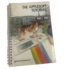 May include: A spiral-bound instructional book titled THE APPLESOFT TUTORIAL with a white cover featuring a vintage illustration of people using early Apple computers, a keyboard, and a rainbow stripe graphic at the bottom with the apple computer inc. logo.