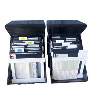 May include: Two black plastic storage cases for 3.5-inch floppy disks, each featuring a hinged lid and a white front panel. The cases are filled with multiple black floppy disks, many with handwritten labels, and include sheets of blank white adhesive labels.