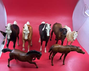 May include: A collection of seven horse figurines in various poses and colors, including dark brown, light brown, black, and white with spots. The horses are arranged in two rows, with some standing upright and others in a dynamic running position.