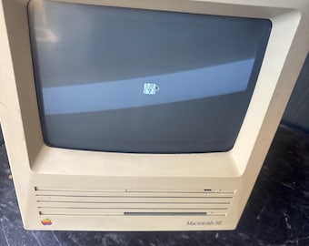 May include: A vintage beige Macintosh SE computer featuring a built-in monitor displaying a floppy disk icon with a question mark. The front panel includes a horizontal disk drive slot, a multi-colored Apple logo, and the text Macintosh SE.