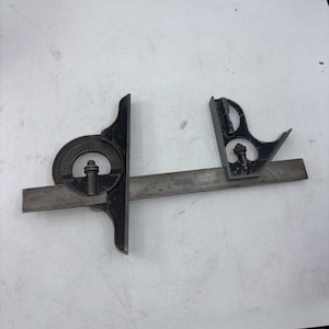 May include: A vintage metal combination square tool featuring a long steel ruler blade with a sliding protractor head and a square head attachment. The components are finished in a dark, weathered black coating with visible metallic silver edges.