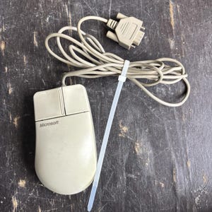 May include: A beige Microsoft computer mouse with two buttons and a long cord ending in a serial connector. The cord is bundled together with a white plastic zip tie. The mouse features a smooth, rounded design with the Microsoft logo printed on it.