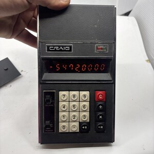 May include: A vintage Craig electronic calculator with a dark grey textured casing and a red LED display showing the number -5472.0000. It features a grid of off-white numeric keys, black function keys, a red clear button, and two vertical switches.