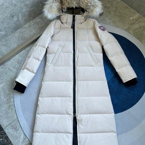 Peut inclure: Un long manteau doudoune couleur crème doté d'une capuche avec une bordure en fourrure épaisse et duveteuse brune et blanche. Le manteau présente une fermeture éclair noire sur toute la longueur, deux poches latérales et des poignets noirs.