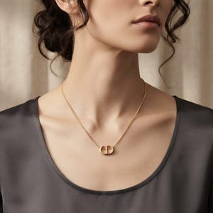 May include: A gold-toned necklace featuring a delicate chain and a central pendant shaped like the letters CD. The pendant has a smooth, metallic finish and is suspended from a fine, linked chain that rests against the collarbone area.