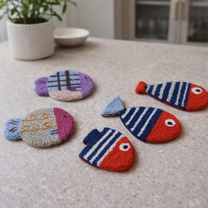 May include: Four punch needle coasters shaped like fish. One is purple with a grid pattern, one is gray with orange swirls, and two are orange and blue with white stripes. Each fish has a distinct tail and eye detail, crafted with textured yarn.