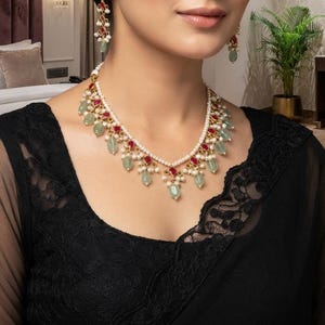 May include: A gold-toned necklace and matching drop earrings set featuring small white pearls, faceted red stones, and pale green teardrop-shaped stones. The jewelry is arranged in a delicate, repeating pattern with gold metal accents and fine chains.