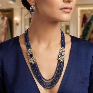Può includere: Una collana a più fili con perline sfaccettate blu scuro disposte su cinque strati, impreziosita da due connettori in metallo dorato con pietre blu esagonali e cristalli trasparenti, abbinata a orecchini pendenti nello stesso stile.