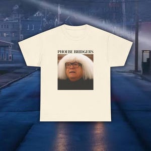 Pode incluir: Uma t-shirt de manga curta cor de creme com um gráfico retangular centrado. O gráfico mostra uma pessoa com uma peruca branca e óculos, com o texto PHOEBE BRIDGERS impresso numa fonte serifada preta diretamente acima da imagem.